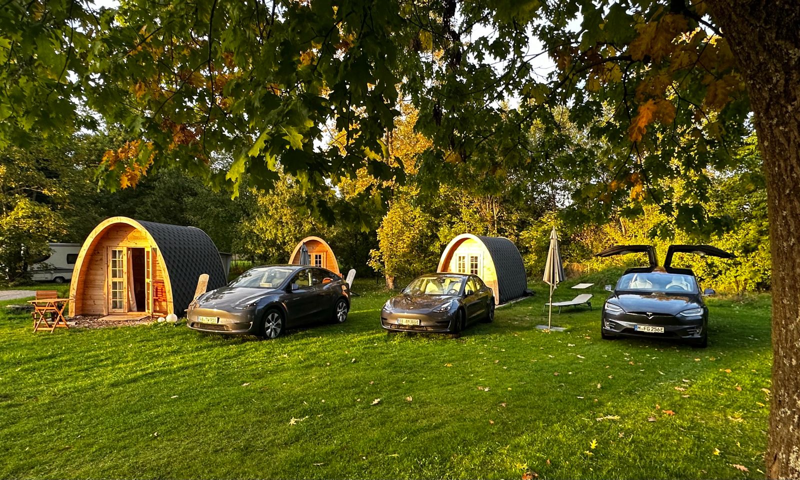 drei Teslas stehen auf grüner Wiese vor drei CampingPods, das sind tonnenförmige Holzhütten des Campingplatzes, die zu mieten sind